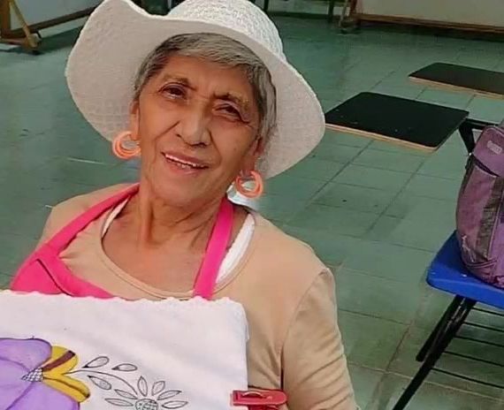 Una señora con sombrero sonríe mientras pinta un mandil en un taller.