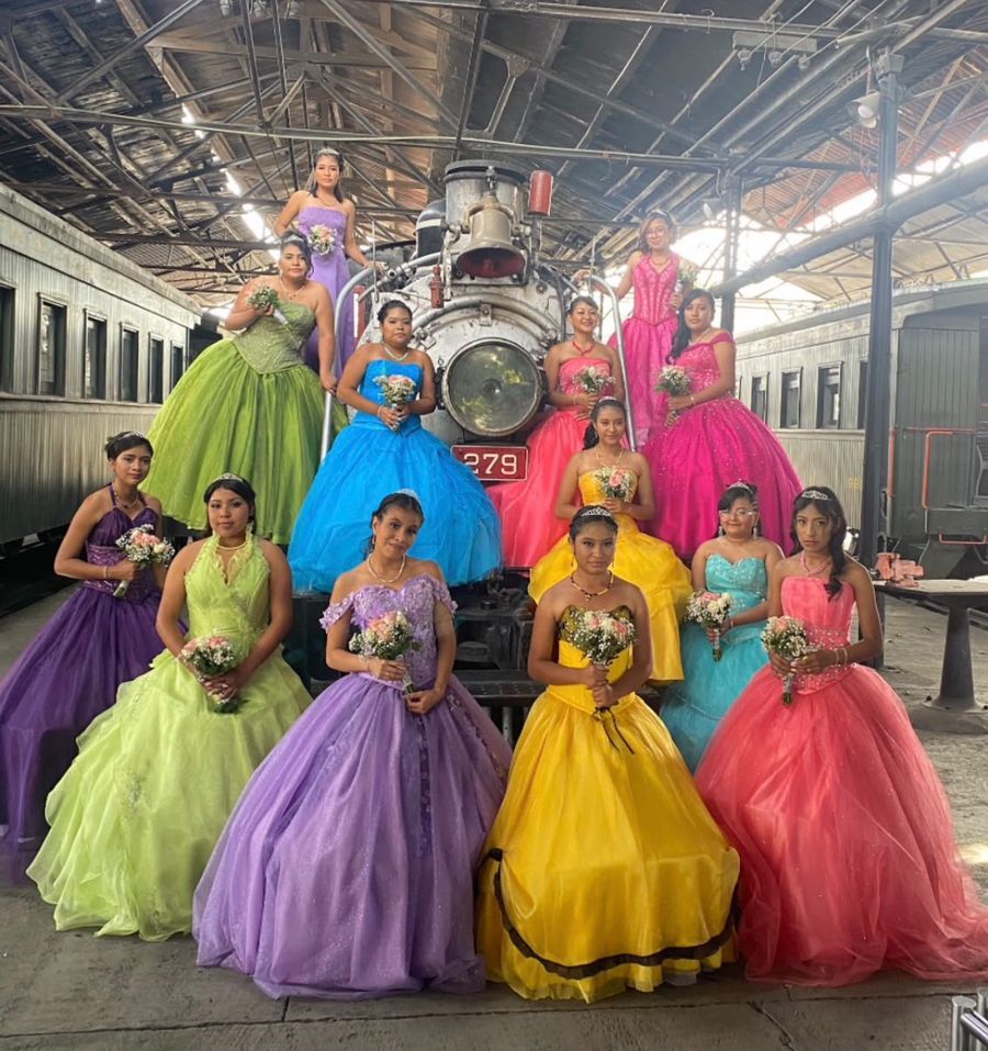 Doce quinceañeras con vestidos de colores y ramos de flores posan frente a una locomotora antigua.